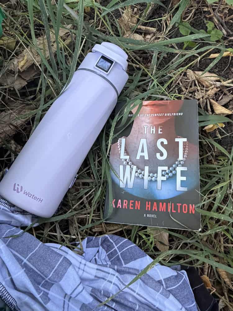 WaterH smart water bottle lying in the grass beside a book called "The Last Wife".